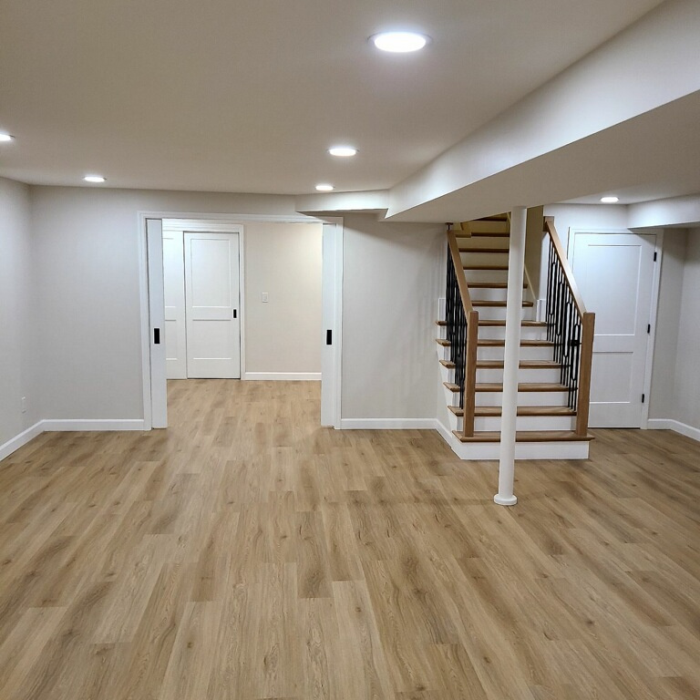 Newly remodeled home basement with wooden flooring, white walls, recessed lighting, and a staircase.